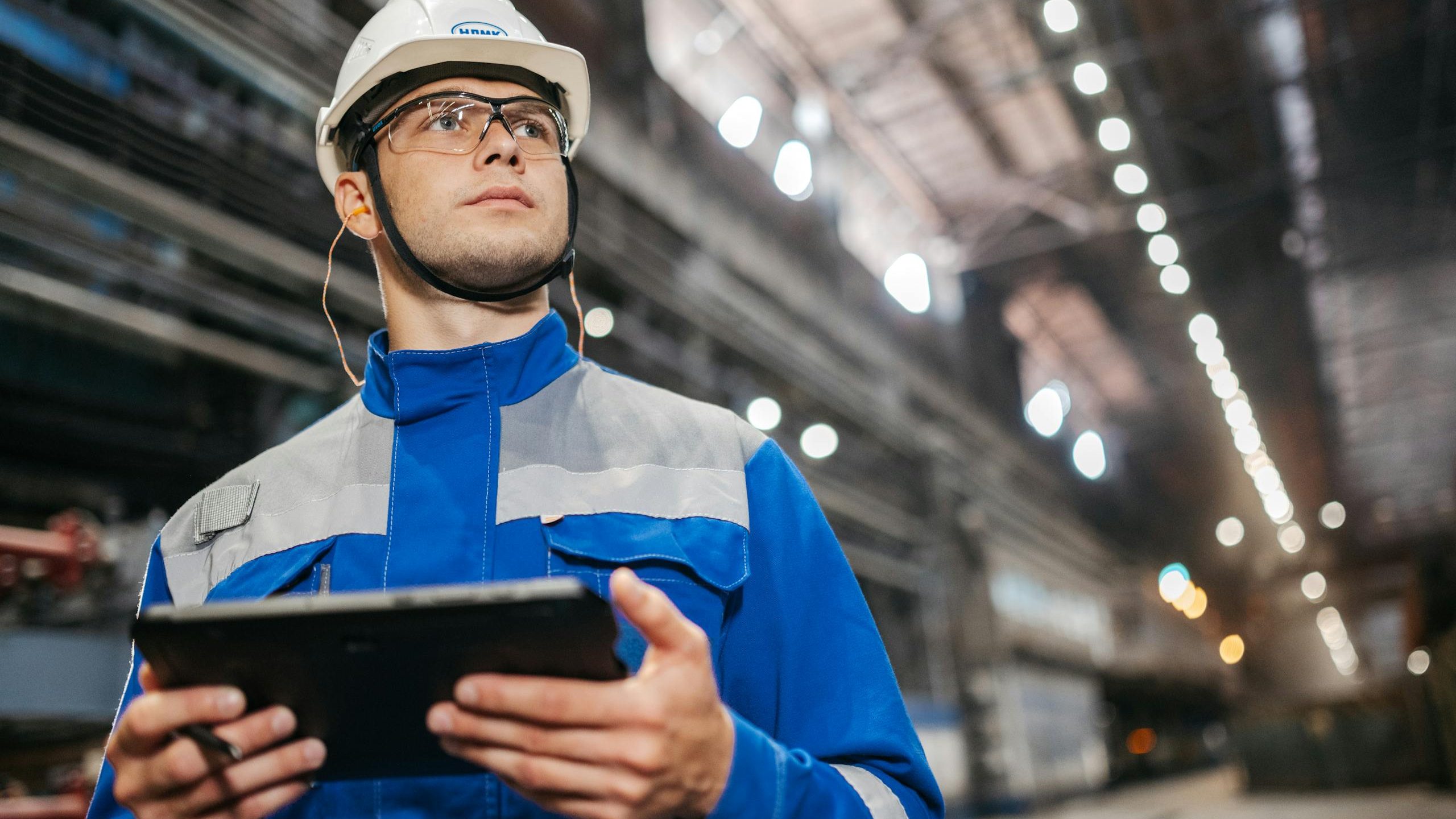 Factory worker in safety gear using a tablet in a well-lit industrial setting.